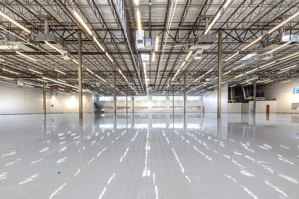 Wide empty commercial floor finished in a solid light grey coating, painted columns and a roll-up door along the back wall