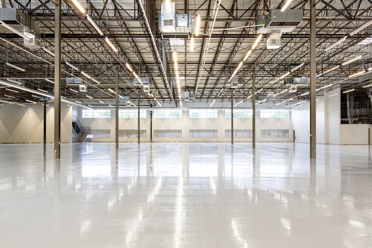 Empty warehouse interior with a high-gloss light grey floor reflecting rows of overhead strip lights and steel roof trusses, clerestory windows along the far wall