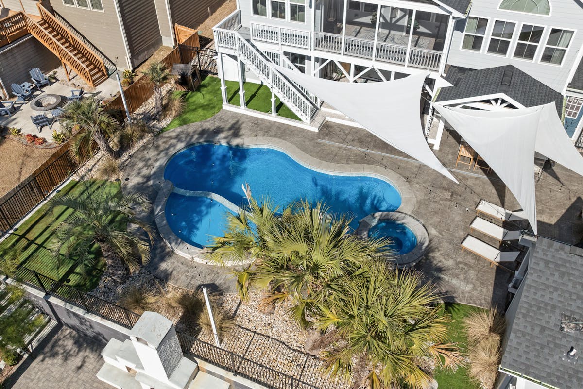 Overhead view of a freeform blue pool ringed by a broad textured concrete deck, white shade sails and palms above it, a screened porch and stairs along the back of the house